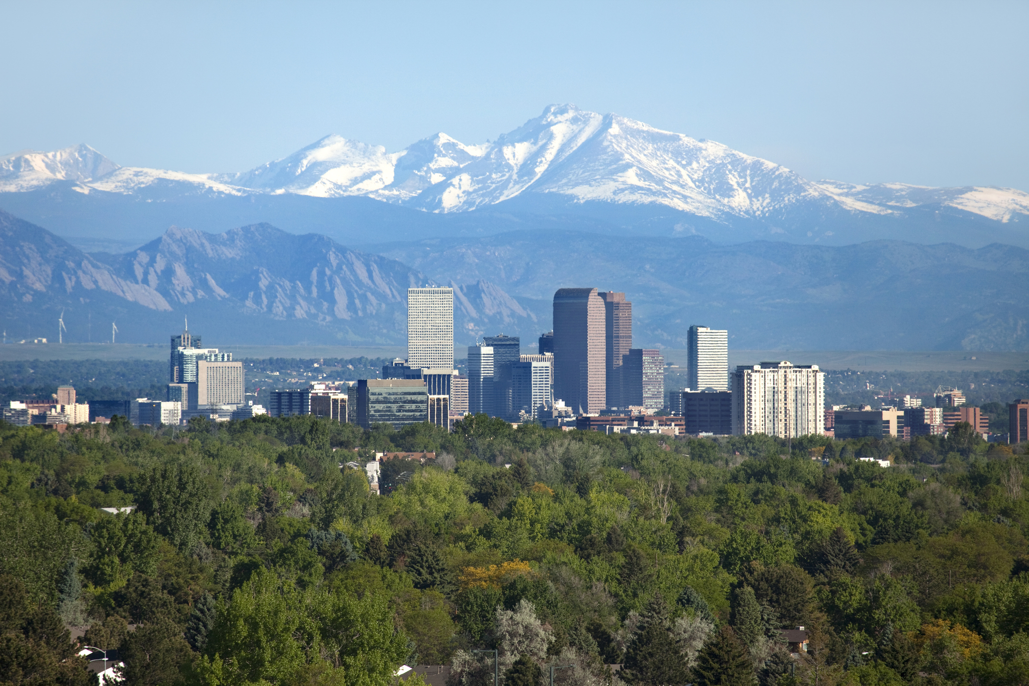 Snow covered Longs Peak, part of the Rocky Mountains stands tall in the background with green trees and the Downtown Denver skyscrapers as well as hotels, office buildings and apartment buildings filling the skyline.